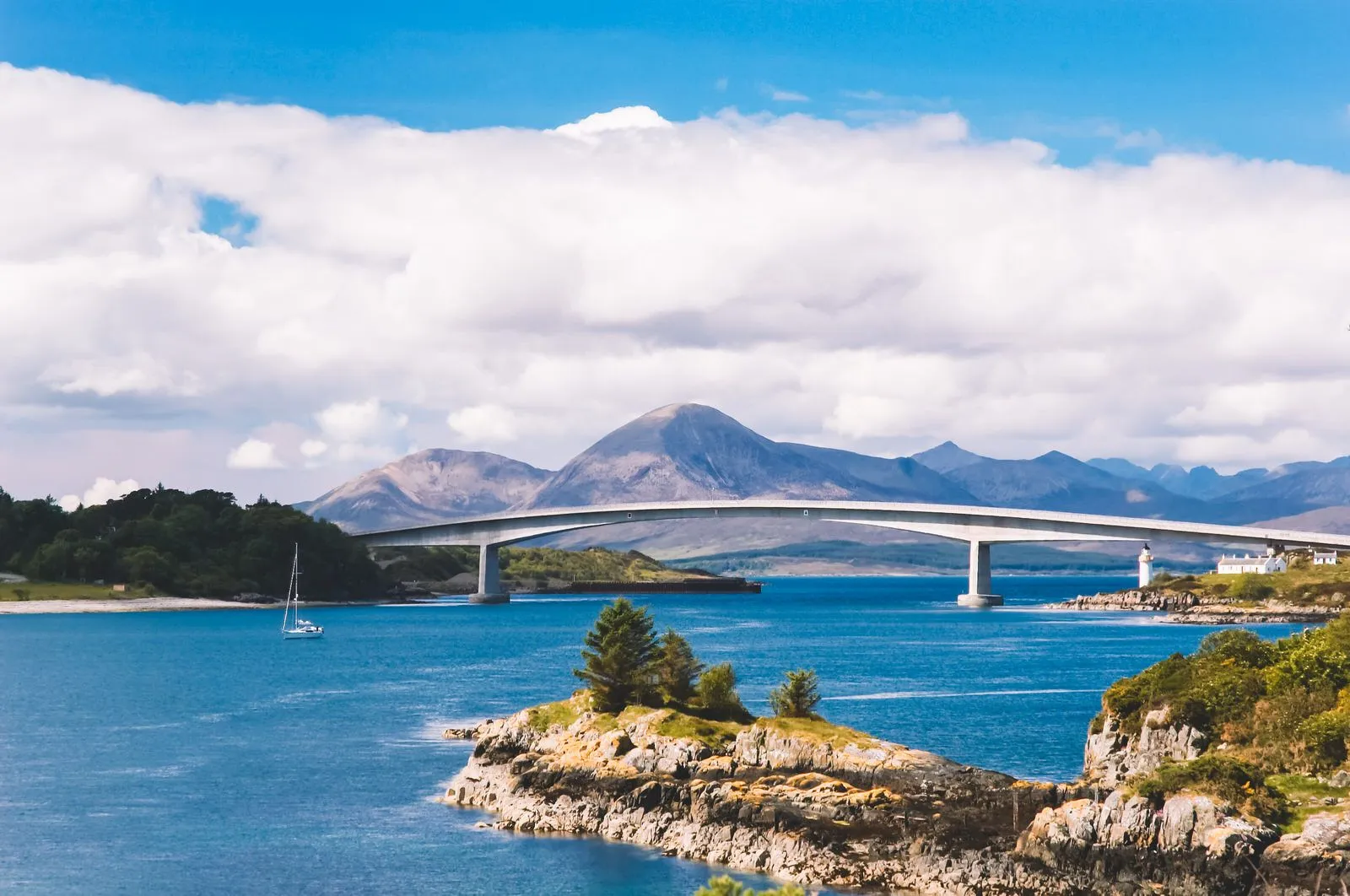 A scenic long shot of a modern bridge spanning across a calm blue body of water, with a small rocky island in the foreground and majestic mountains under a cloudy sky in the background. A single sailboat is visible near the shoreline on the left.