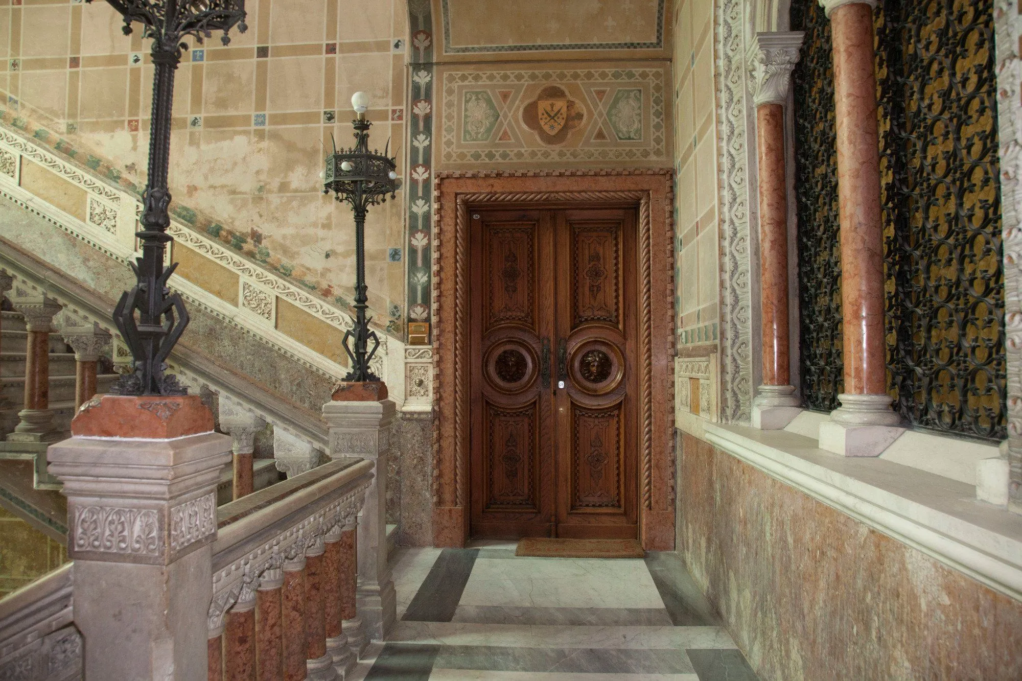 An ornate interior featuring a richly carved wooden door, marble staircase, and decorative walls. The composition is well-balanced, drawing attention to the door and showcasing the intricate details of the architecture.