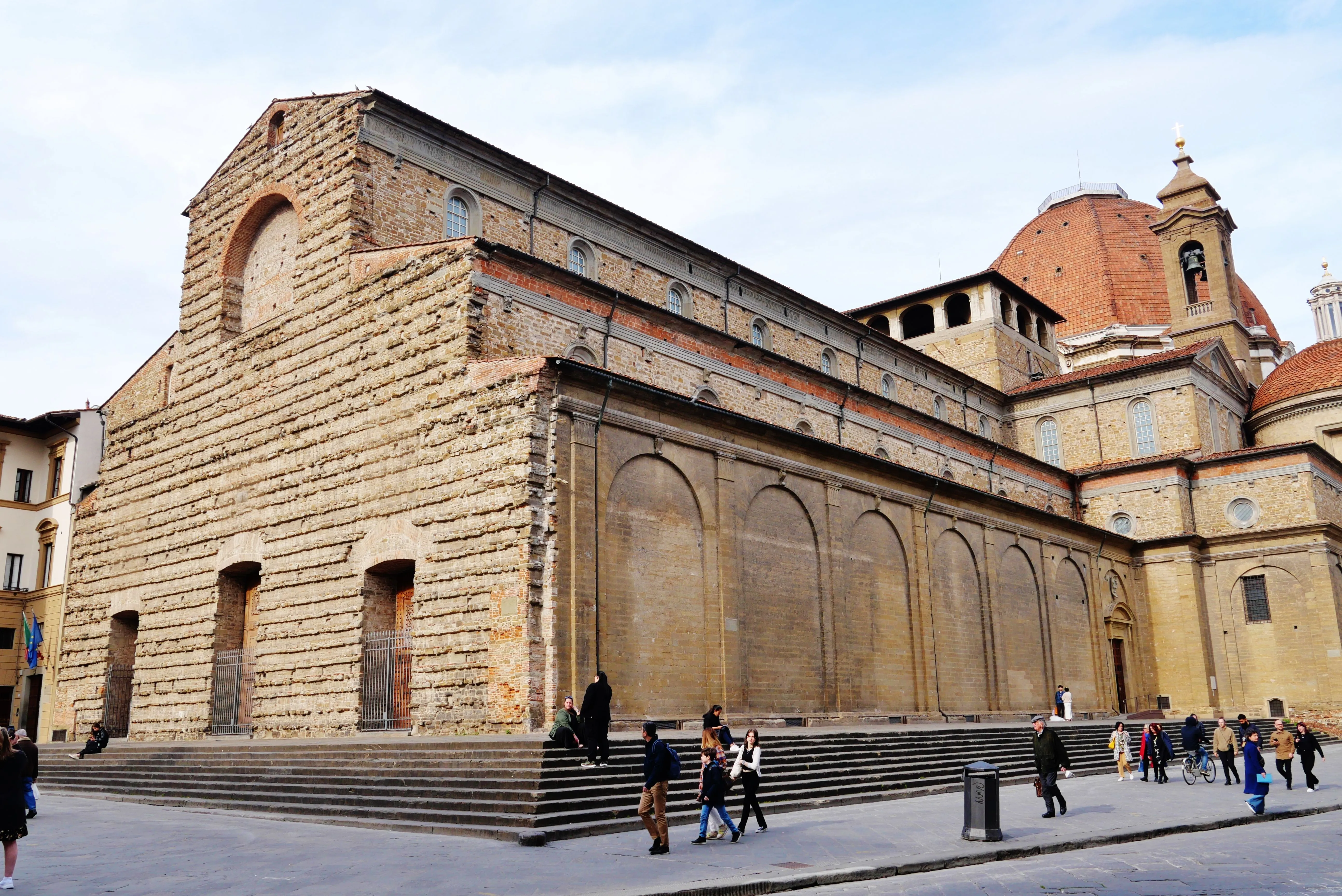An editorial-style image capturing the side of a large, historic stone building with prominent steps, featuring several people walking and sitting in the foreground under natural lighting.
