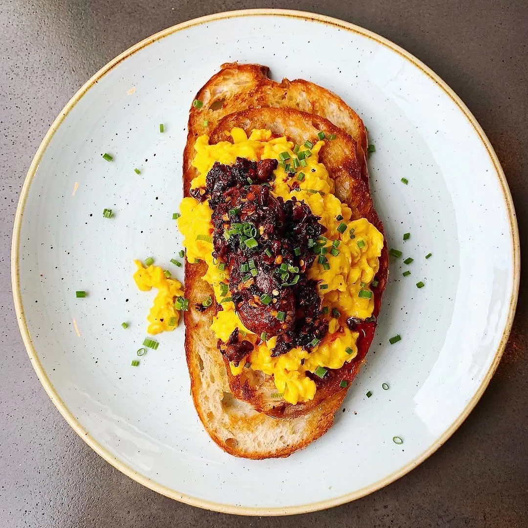 The image captures a well-presented dish of scrambled eggs on toasted bread, topped with a dark savory condiment and fresh chives, served on a light-colored plate.
