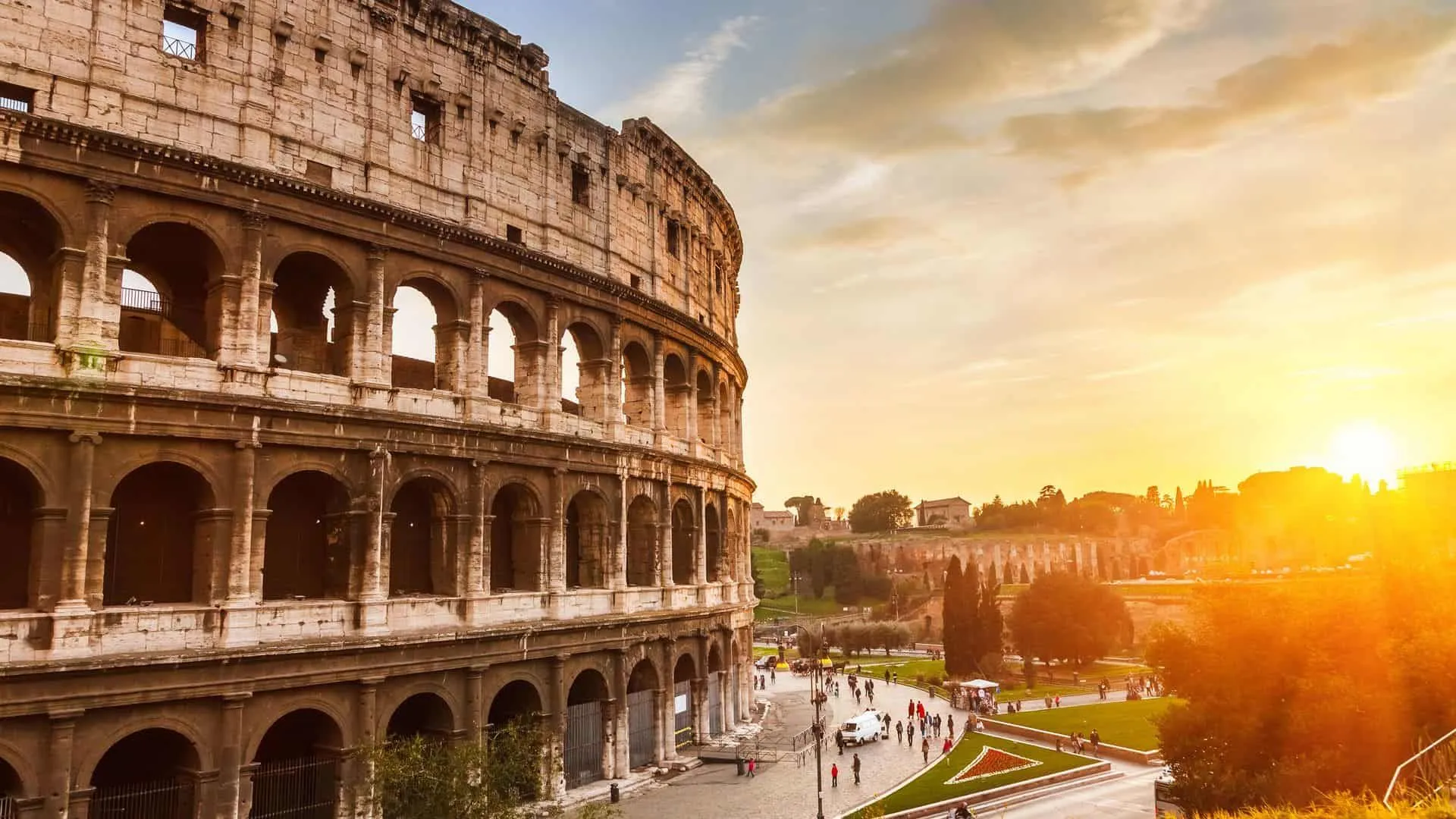 An iconic image of the Colosseum in Rome during sunset or sunrise, showcasing its grandeur. The warm light bathes the ancient structure, highlighting its details against a vibrant sky with scattered clouds. People are visible in the foreground, providing a sense of scale and activity.