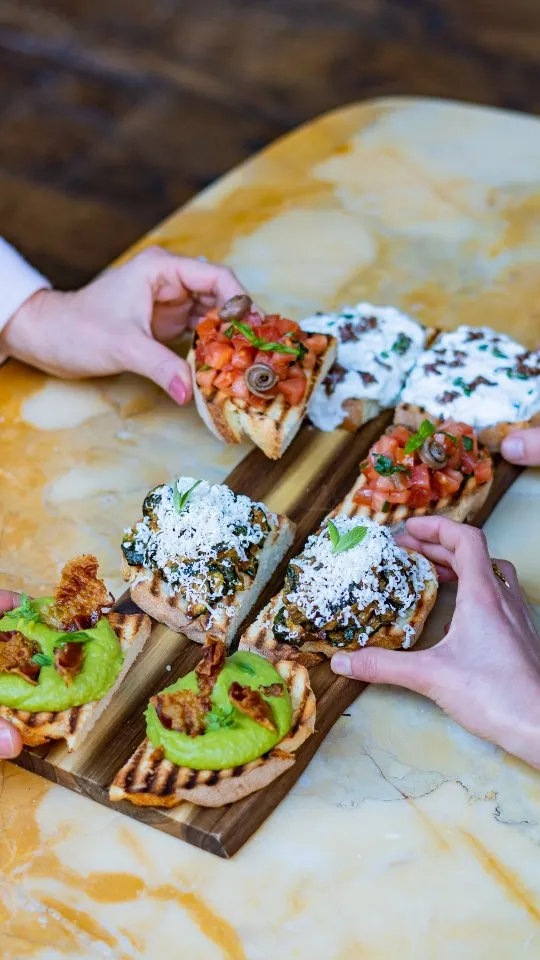 An inviting shot of various gourmet bruschetta or crostini served on a wooden board, with multiple hands reaching for the pieces, conveying a sense of sharing and community dining. The food is vibrant and appealing.
