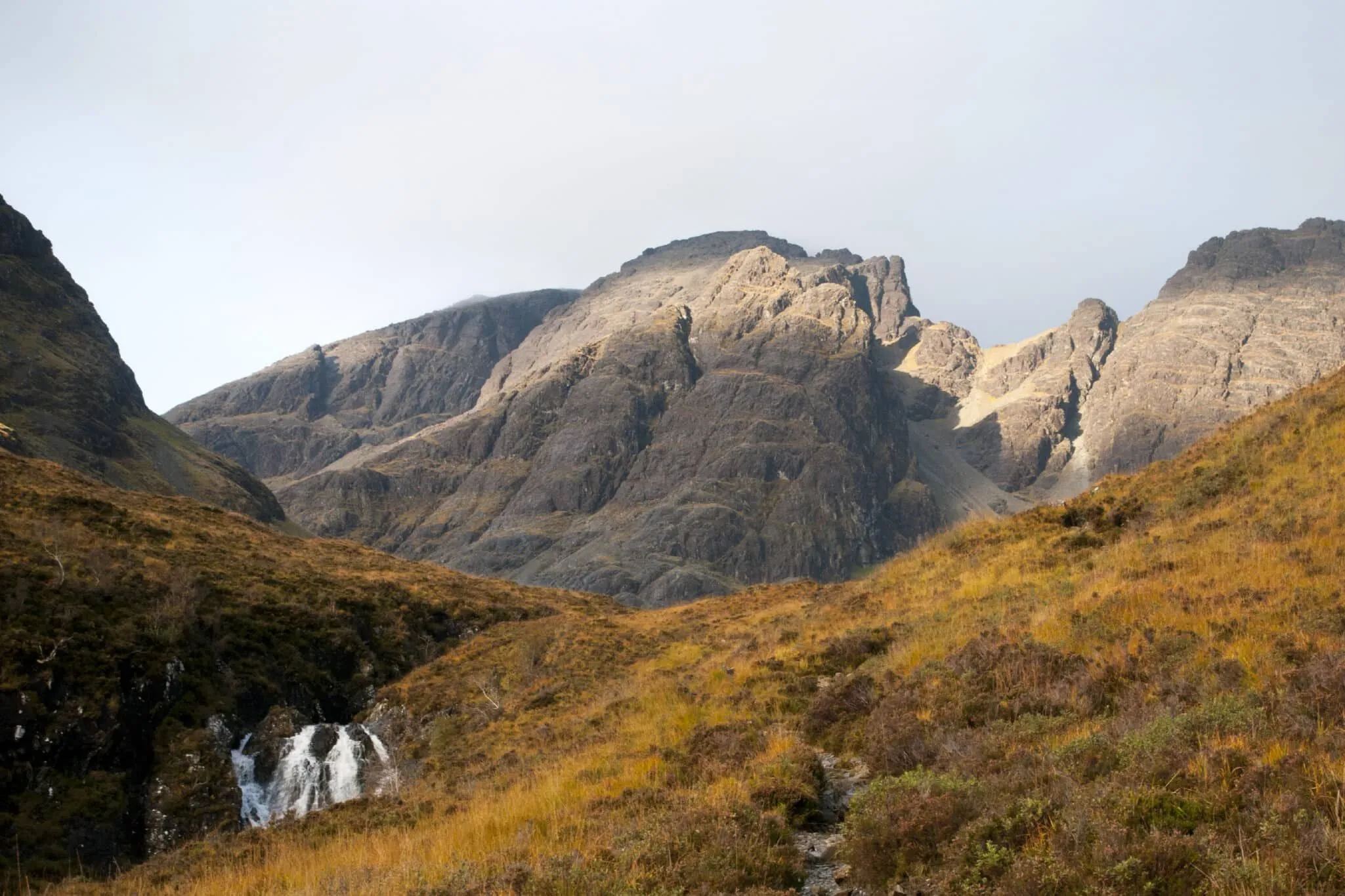 A rugged mountain landscape with a small waterfall in the foreground and a vast, rocky mountain range under a bright sky. The terrain is covered in sparse, yellowish-brown vegetation.