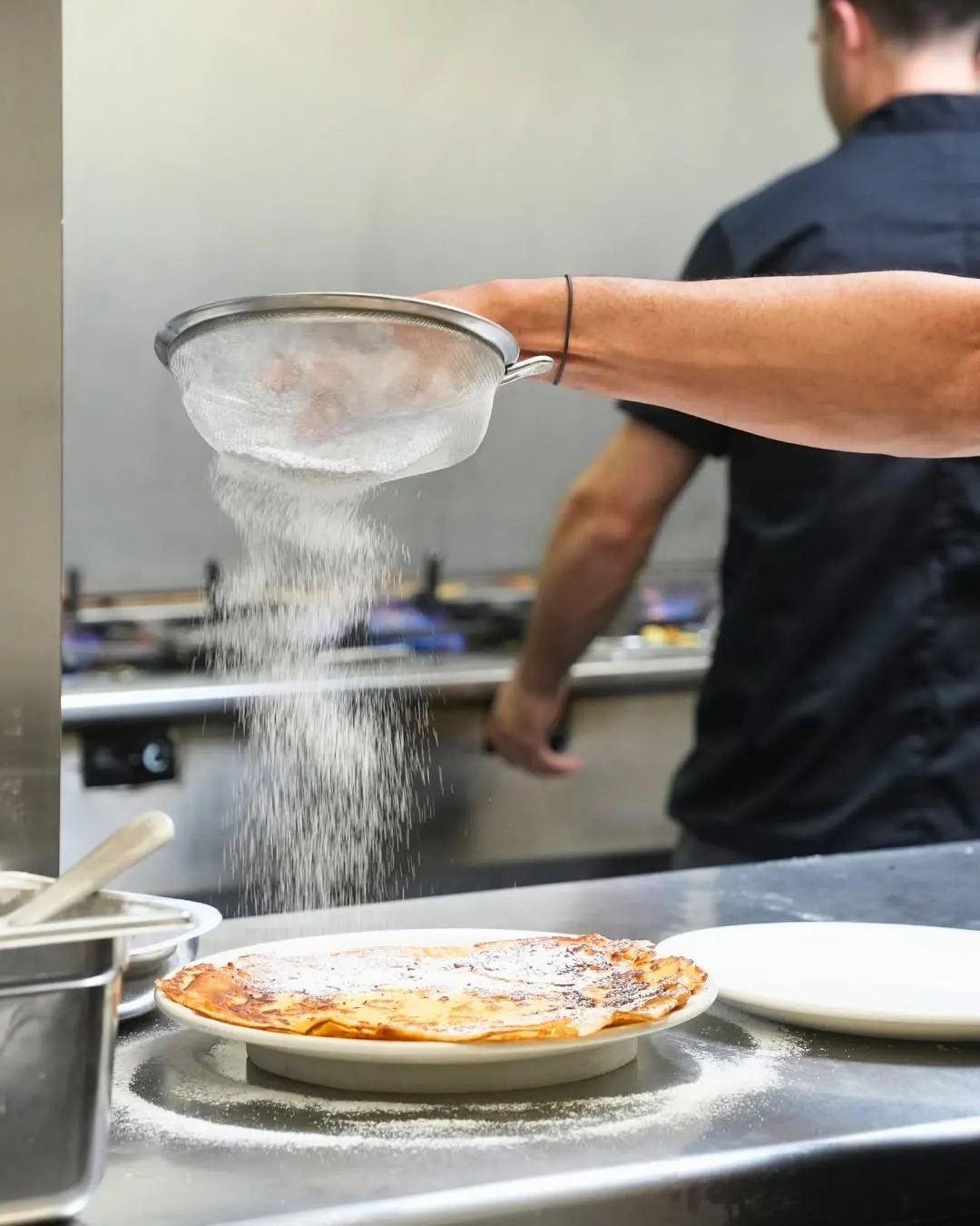A close-up shot capturing a chef's hand sifting powdered sugar over a stack of crepes in a professional kitchen environment. The falling sugar creates a dynamic visual element. The background is softly blurred, keeping the focus on the food and the action.