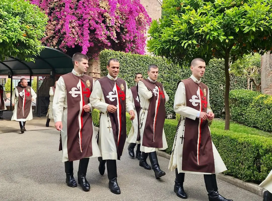 A group of individuals in distinctive traditional garments walk along a garden path with lush greenery and vibrant purple bougainvillea.