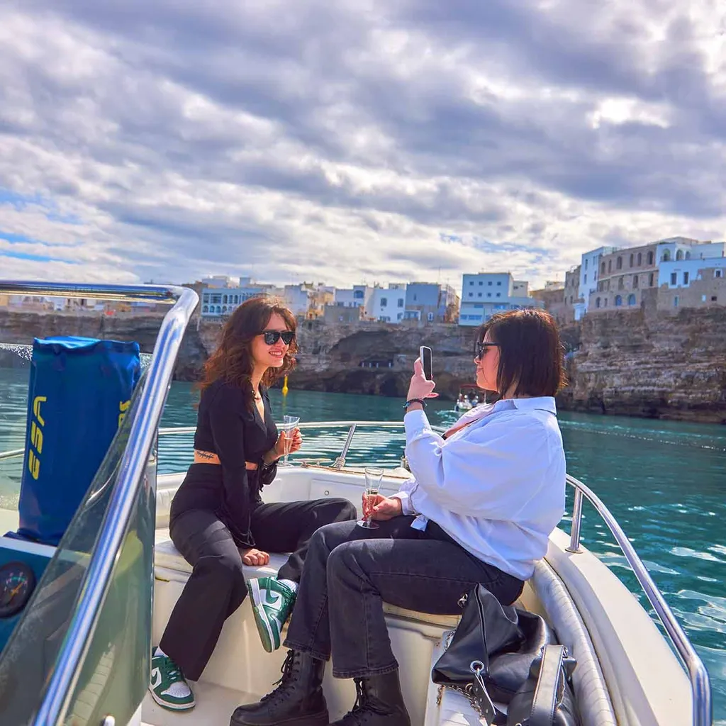 An image capturing two women on a boat enjoying the view of a coastal town, with one person taking a photo of the other. The scene is bright and shows clear blue water and a cloudy sky.