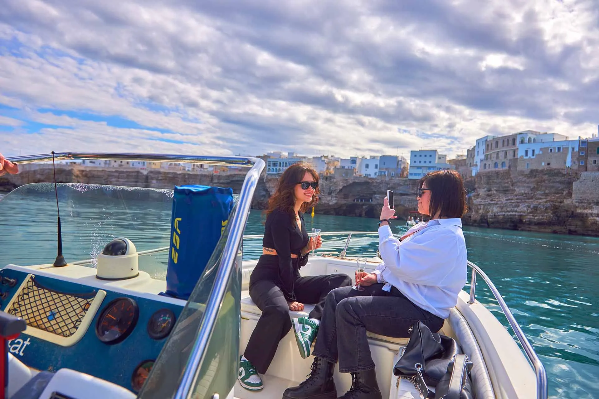 An image capturing two women on a boat enjoying the view of a coastal town, with one person taking a photo of the other. The scene is bright and shows clear blue water and a cloudy sky.