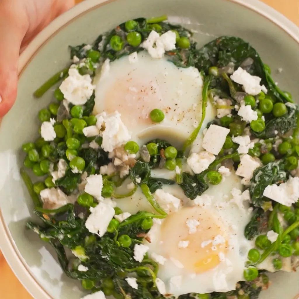 A close-up, top-down view of a savory breakfast dish featuring two eggs cooked in a bed of green spinach and peas, topped with crumbled white feta cheese. The dish is presented in a light-colored bowl.