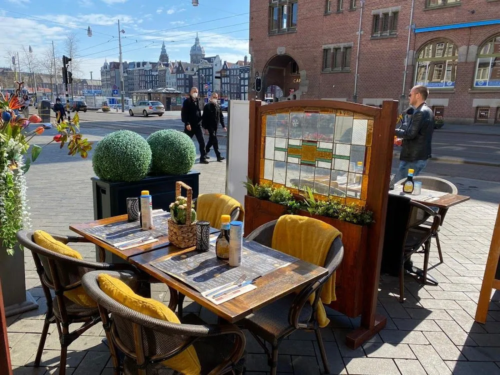 An inviting outdoor cafe terrace on a sunny day, with two tables set with menus, condiments, and yellow accent blankets and cushions. In the mid-ground, pedestrians walk on the street, and historical city buildings are visible in the background under a clear blue sky.