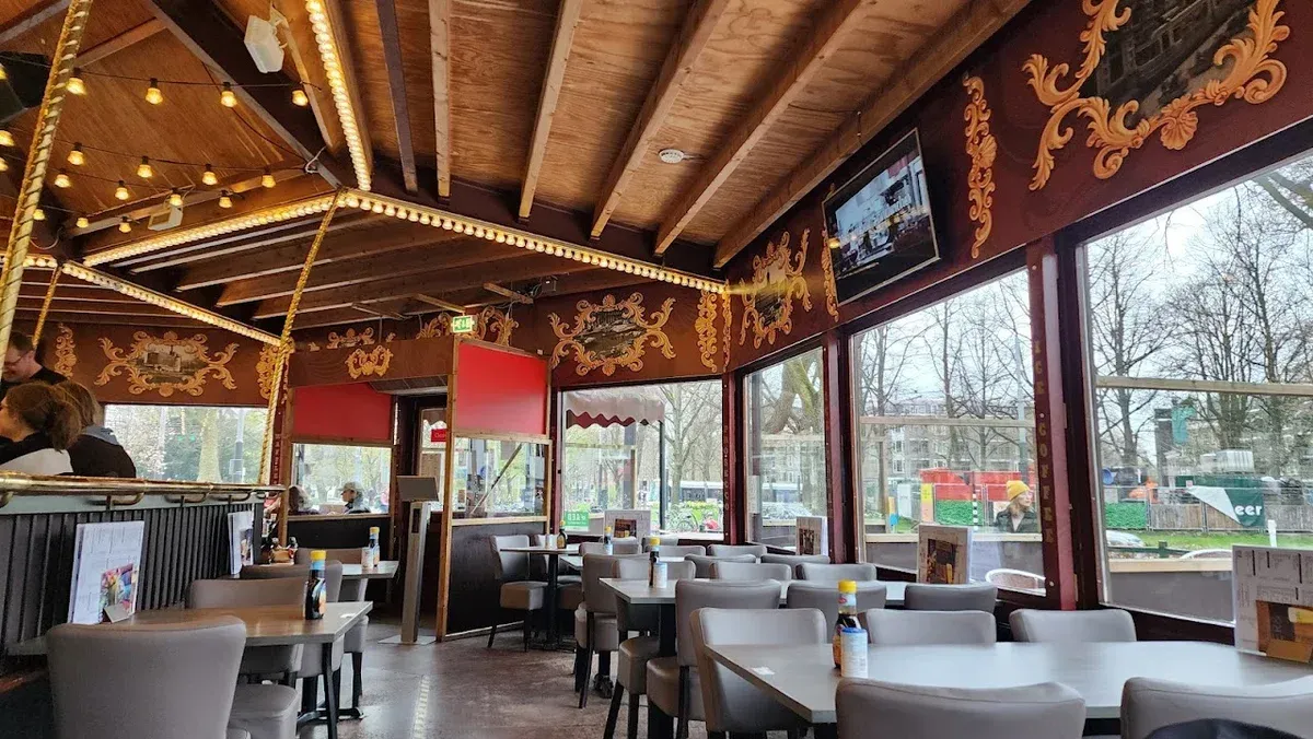 An inviting interior shot of a restaurant, featuring a warm wooden ceiling with string lights, decorative red and gold walls, and neatly arranged grey tables and chairs. Large windows offer a view of the outside street scene. The space appears clean and well-maintained.