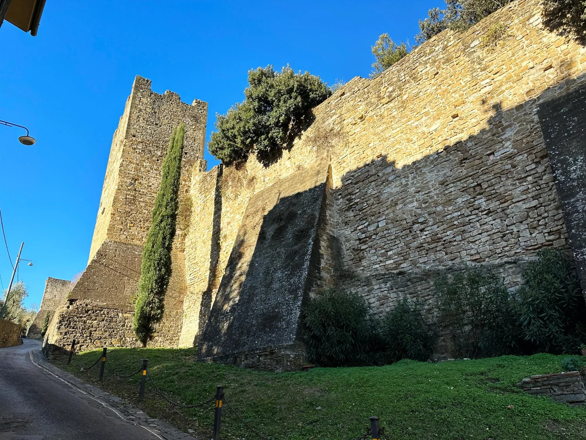 An editorial-style image depicting a towering ancient stone wall and watchtower, with a tall cypress tree standing beside it, set against a clear blue sky. The sunlight casts strong shadows across the wall, highlighting its texture.
