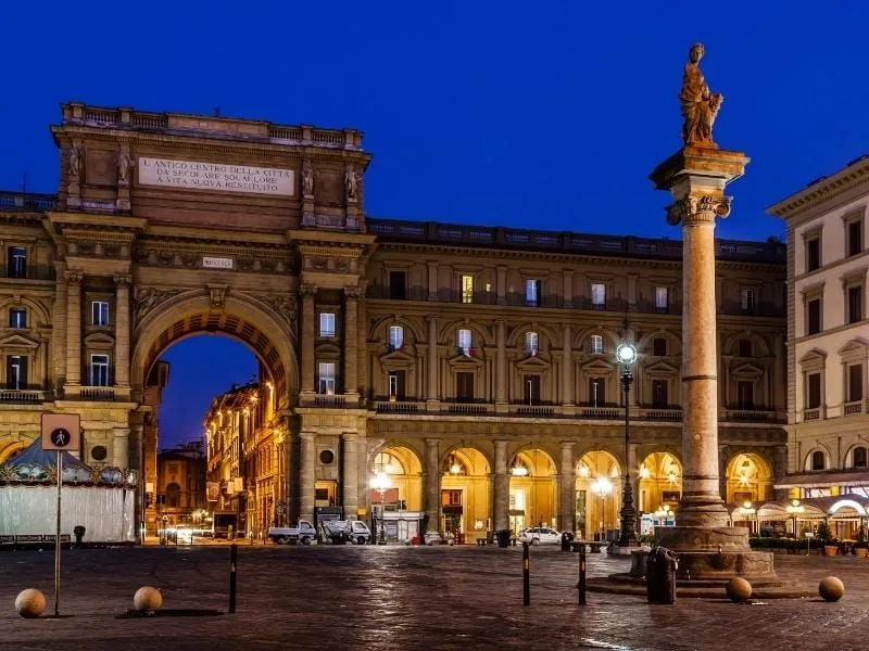 An editorial style image showcasing the Arch of the Triumphs in Piazza della Repubblica, Florence, Italy, at twilight. The historic architecture is well-lit, highlighting details against a deep blue sky.