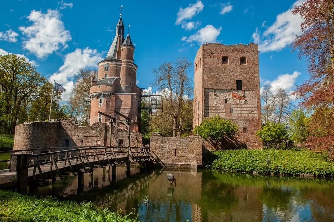 A well-preserved historic brick castle featuring a prominent round tower, a square tower, and a wooden drawbridge spanning a reflective moat under a clear blue sky with white clouds. Lush green foliage surrounds the water.