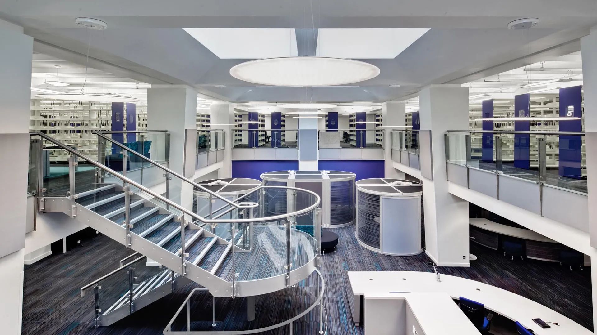 A well-composed and well-lit image of a modern multi-level interior space, possibly a library or a contemporary office, featuring a prominent curved staircase with glass railings, circular seating booths, and an overhead circular light fixture. The color palette is primarily blue, white, and gray, contributing to a clean and sophisticated aesthetic. The background shows additional levels with shelving.