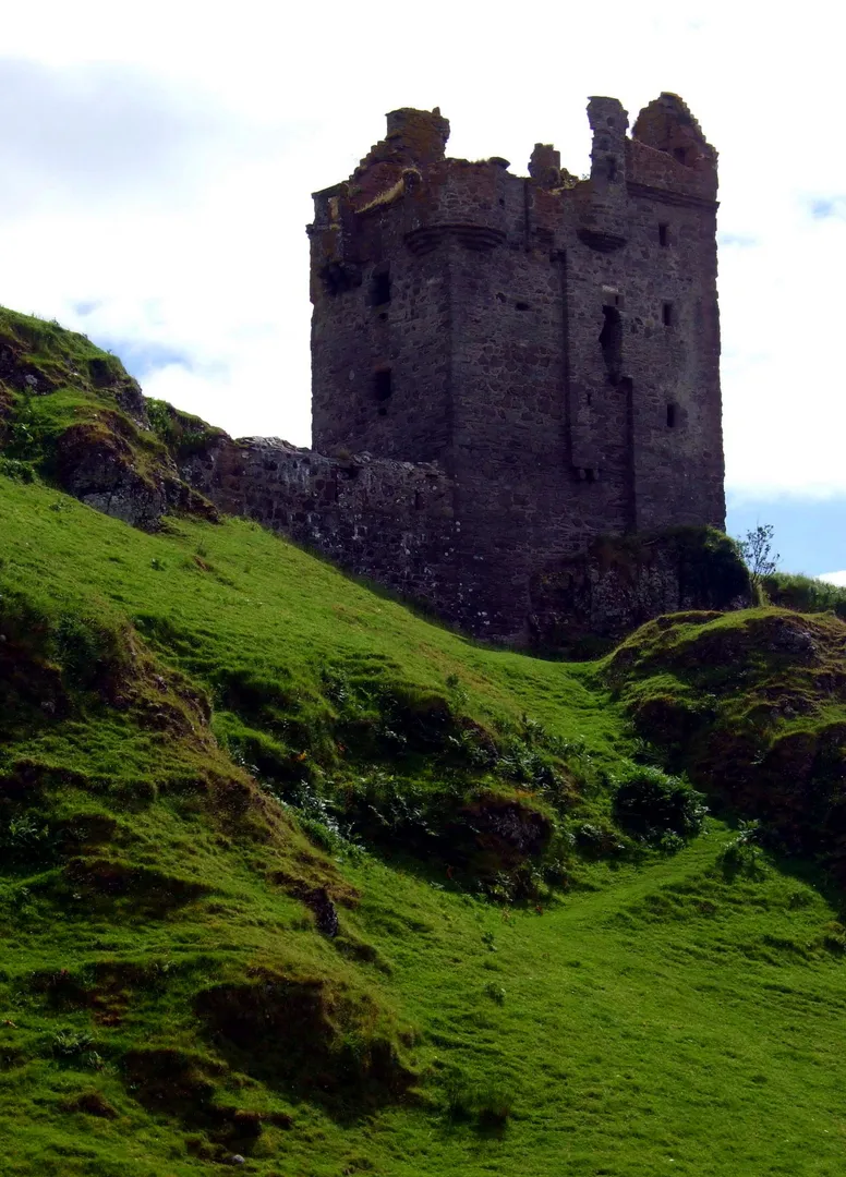 An editorial-quality image featuring a historic stone castle tower perched atop a steep, vibrant green hillside. The sky is partly cloudy, and the overall lighting is good.