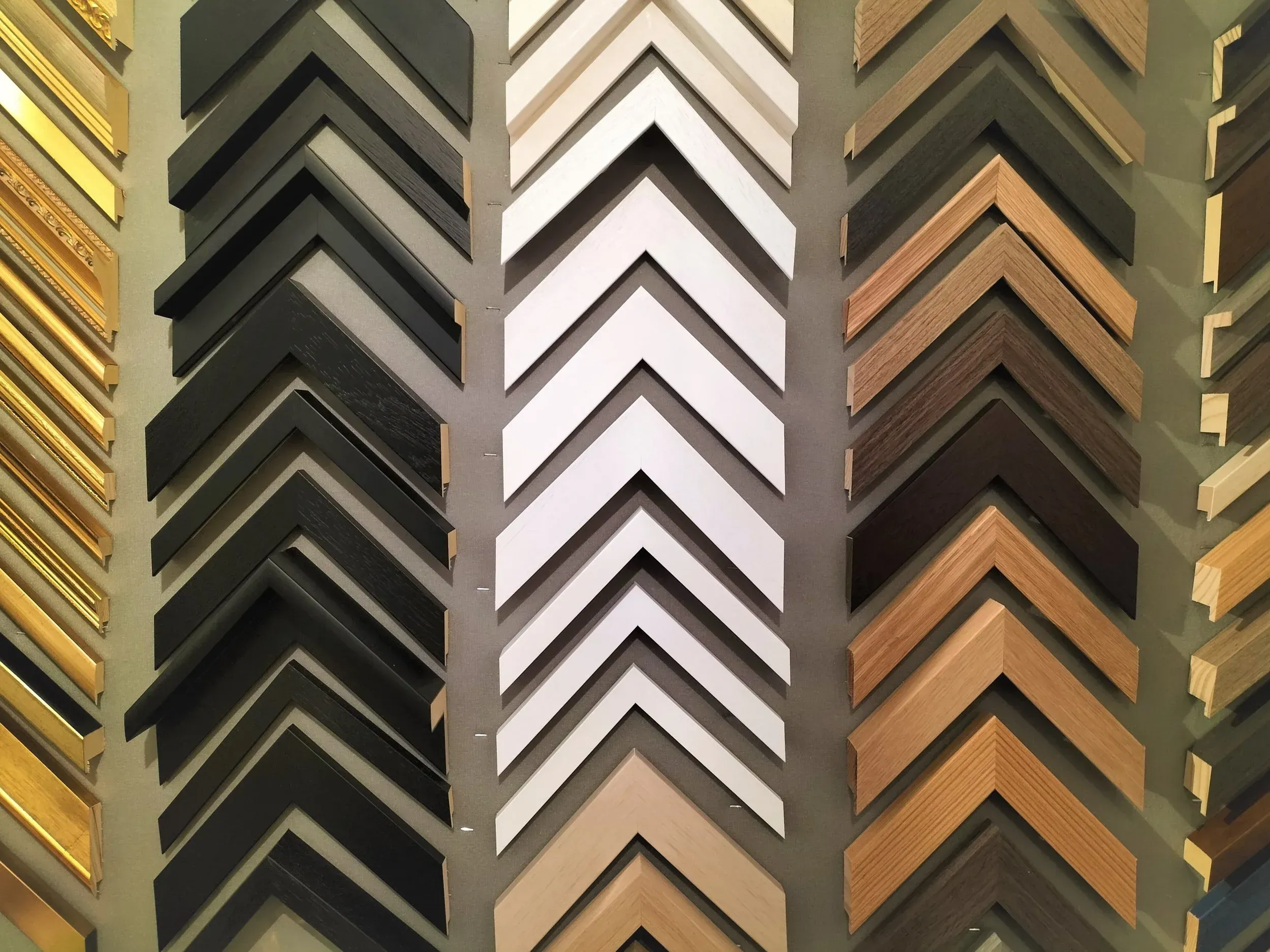 A close-up, high-angle shot showcasing a neatly arranged display of various picture frame corners. The frames are presented in three vertical columns, with samples in black, white, and natural wood tones, each featuring slightly different profiles and finishes. The arrangement emphasizes the variety of options available.