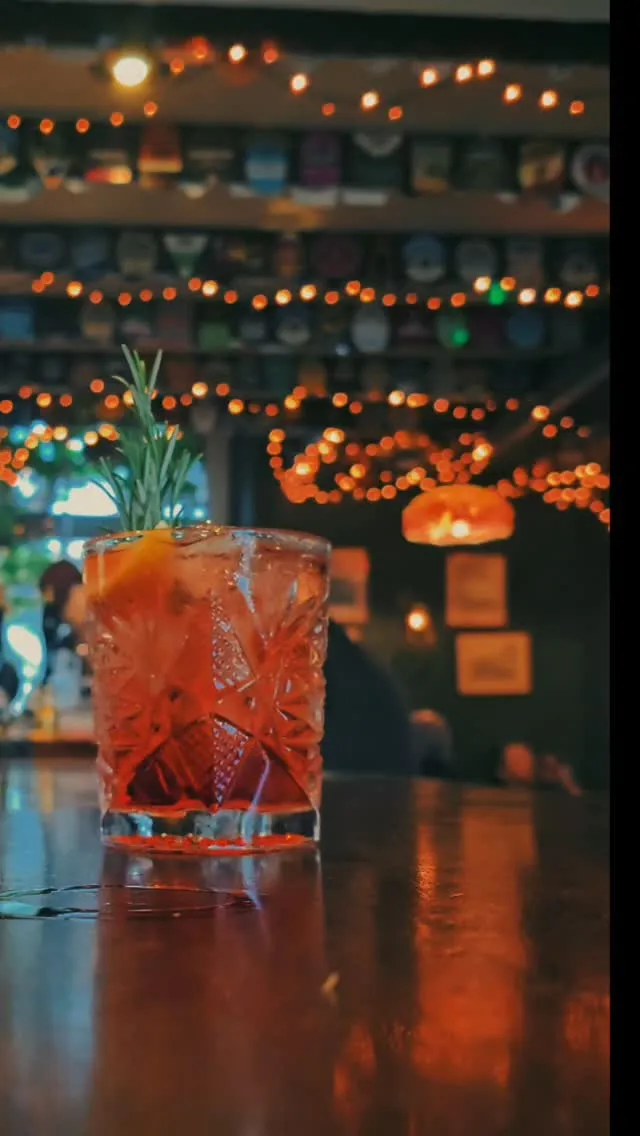 A well-composed and sharp close-up of a cocktail in a decorative glass, garnished with rosemary and a citrus slice. The drink is well-lit, showcasing its vibrant color, against a softly blurred background of warm, atmospheric bokeh lights.