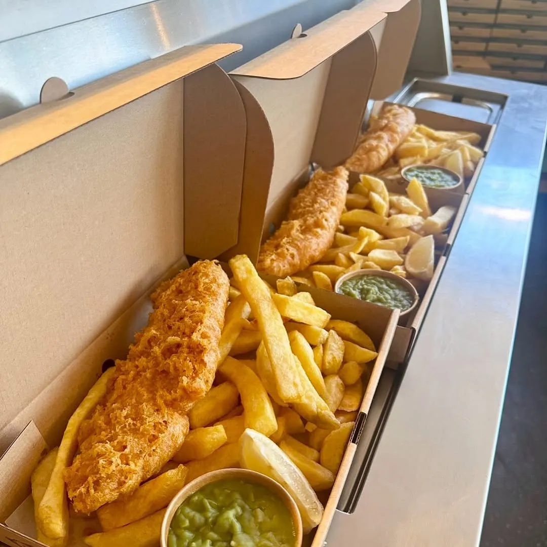 A well-lit and sharp image showcasing multiple servings of fish and chips with mushy peas and lemon wedges in takeaway boxes, appearing photo-realistic with accurate colors.