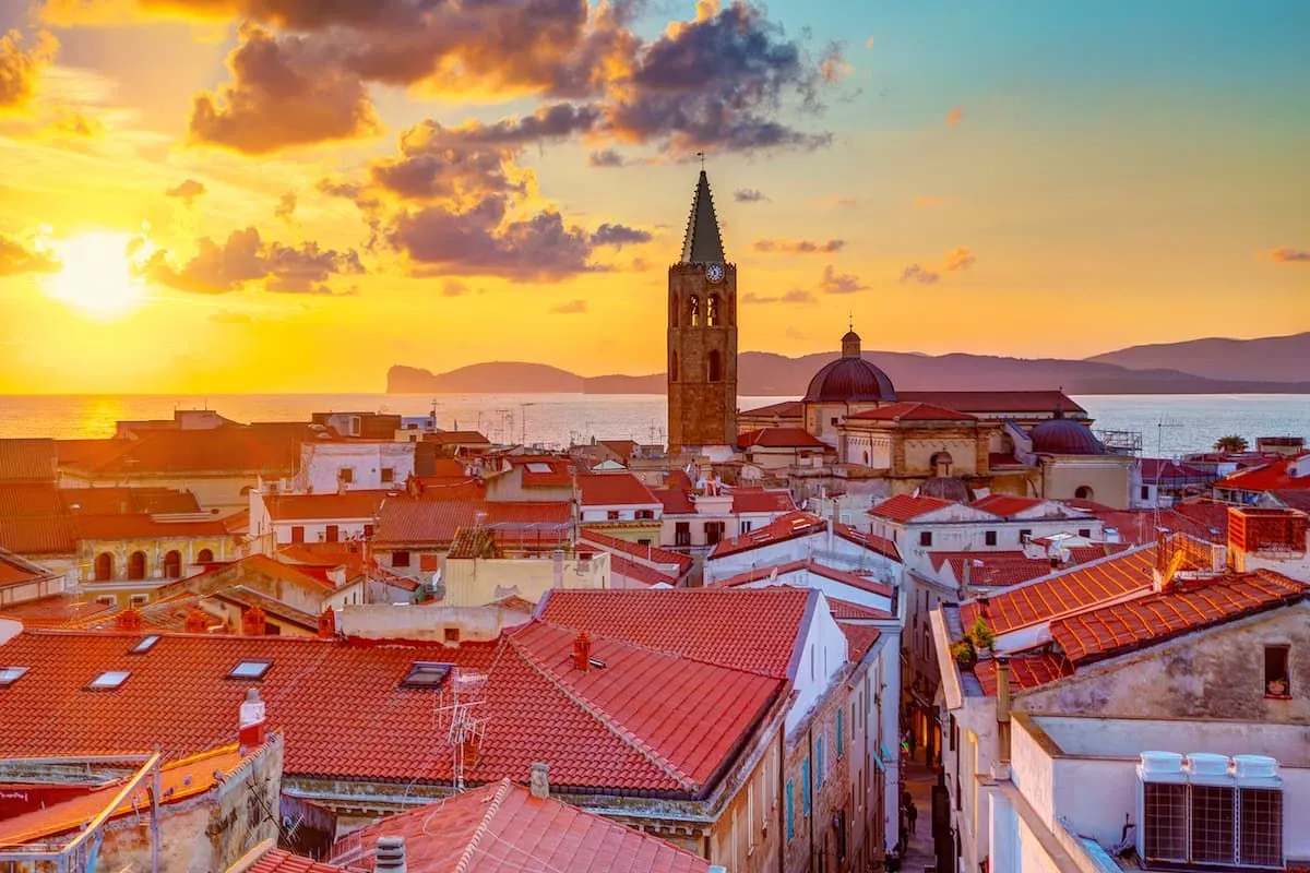 An aerial or elevated view of a coastal city at sunset, featuring traditional red-tiled rooftops, a prominent bell tower, and the sea with distant landmasses under a dramatic orange and blue sky with clouds.