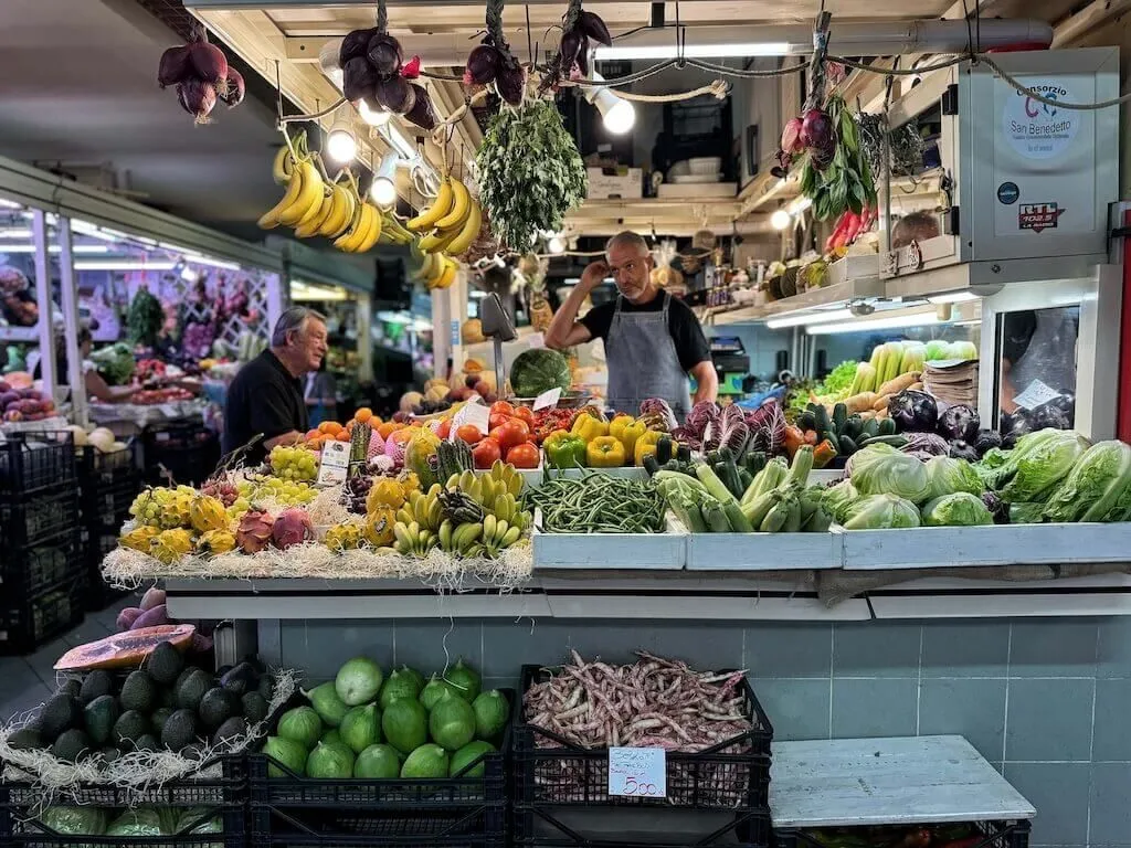 A vibrant and colorful fresh produce market stall, showcasing a wide array of fruits and vegetables, with two market vendors tending to the display. The image captures the authentic atmosphere of a bustling market.
