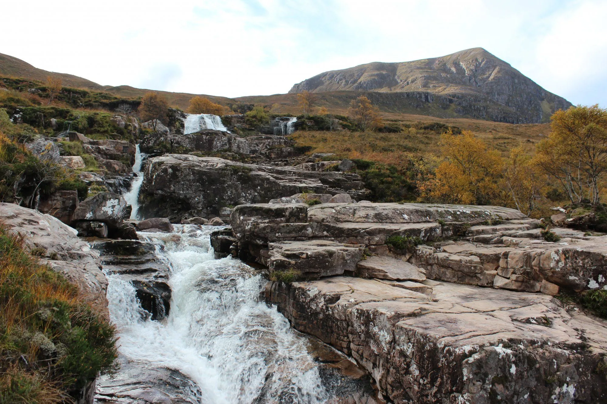 A scenic natural landscape featuring a multi-tiered waterfall cascading over rocky terrain, with a large mountain in the background. The surroundings show autumnal foliage with yellow and brown tones.