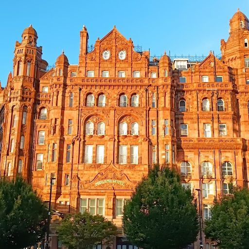 This image is a well-composed exterior shot of a grand hotel. The building is the primary subject and is well-lit, showcasing its intricate details and architectural features. The composition is balanced, with the building taking up most of the frame and trees framing the sides. The image is sharp, capturing the texture of the brick and stone. The colors are accurate and vibrant, reflecting the warm tones of the building and the golden light of the sky. The image is photo-realistic and captures the grandeur of the hotel. The key elements, including the building and its details, are in focus. The image is free of unwanted elements, such as selfies, promotional graphics, advertisements, or cluttered environments. Overall, this is a high-quality image that effectively captures the essence of the hotel's exterior.