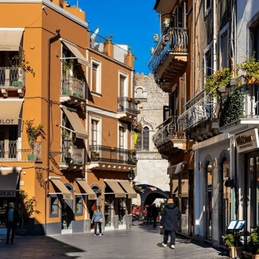 An editorial-style image capturing a narrow, sunlit street lined with multi-story historic buildings in a Mediterranean town. People are walking along the street, and shops are visible at ground level.