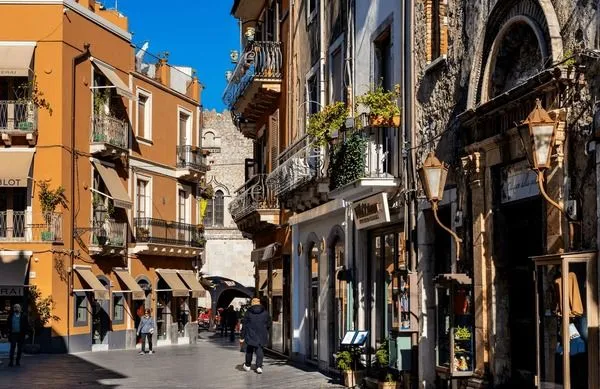 An editorial-style image capturing a narrow, sunlit street lined with multi-story historic buildings in a Mediterranean town. People are walking along the street, and shops are visible at ground level.