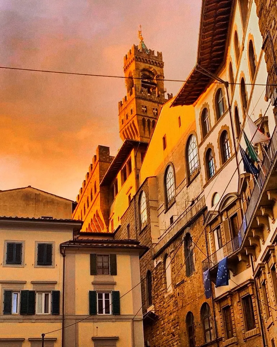An eye-level shot captures a towering, medieval-style stone building against a dramatic, fiery orange sky, suggestive of a strong sunset. Lower buildings with varying architectural styles and shuttered windows flank the main structure, showcasing a street view in an ancient city. Flags hang from some of the balconies on the right.