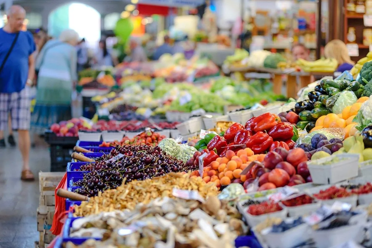 An editorial image showcasing a vibrant array of fresh produce at a market stall. The shallow depth of field effectively blurs the background, focusing on the foreground elements like cherries, red peppers, apricots, and various other fruits and vegetables. The lighting is natural and enhances the rich colors of the produce.