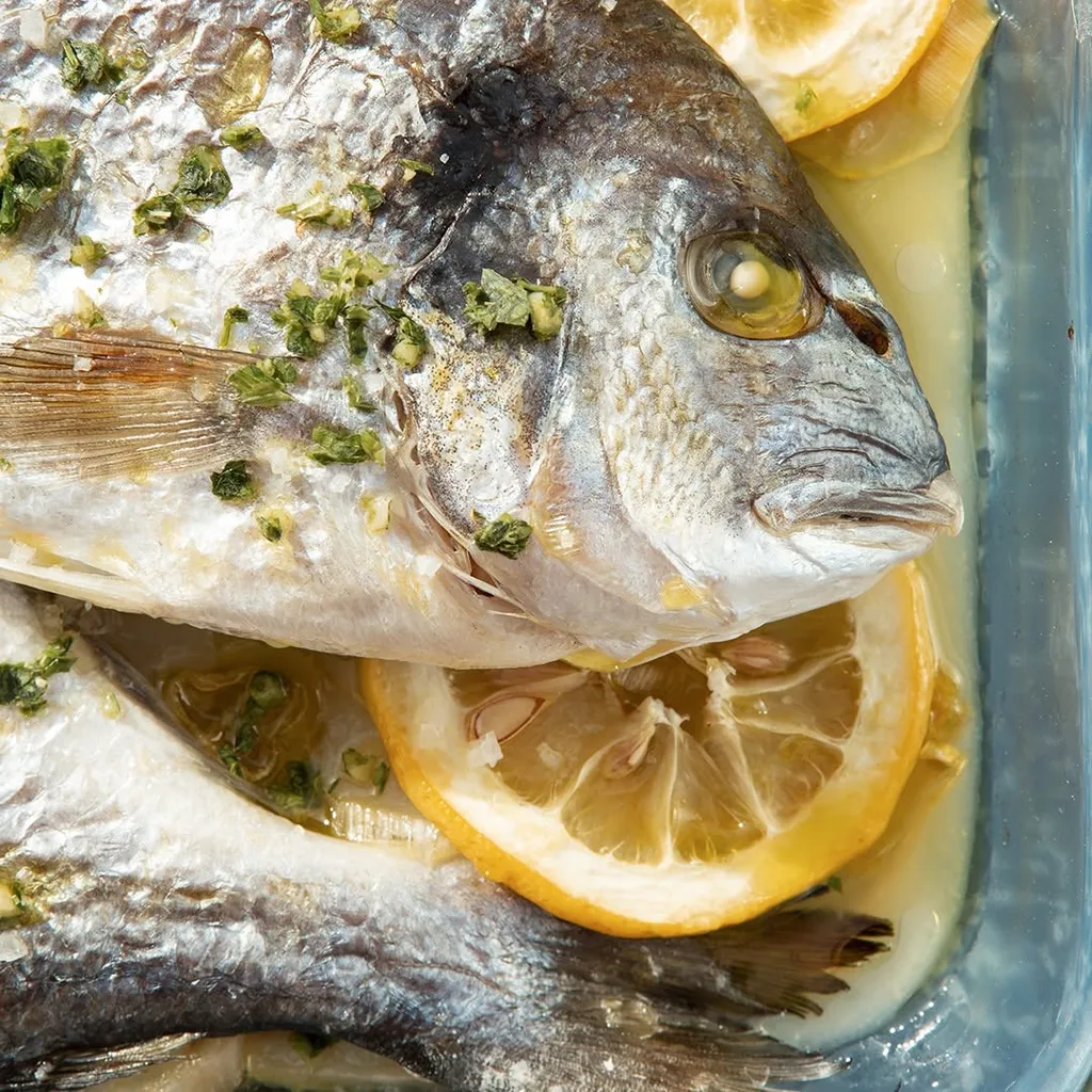 A professionally taken close-up of a cooked whole fish, garnished with green herbs and lemon slices, served in a clear glass baking dish.