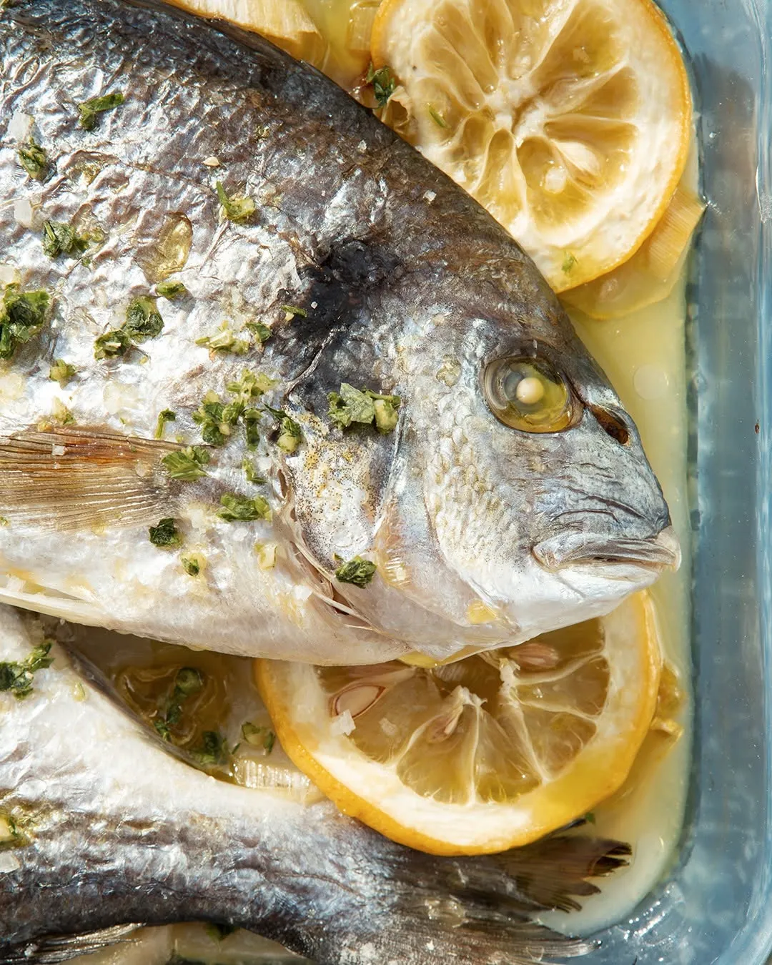 A professionally taken close-up of a cooked whole fish, garnished with green herbs and lemon slices, served in a clear glass baking dish.