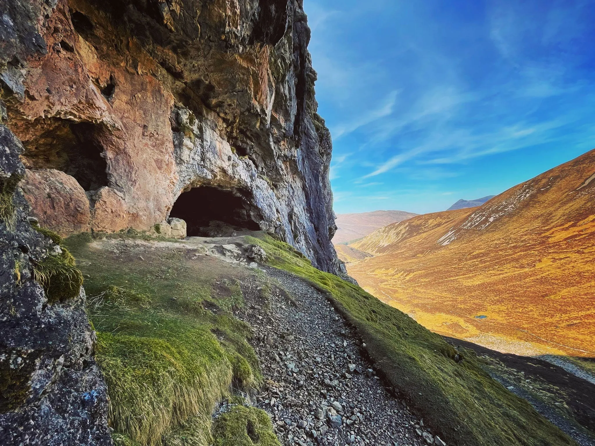 The image is well-composed with strong leading lines from the path and cliff face guiding the eye towards the expansive valley and bright sky. Lighting is generally good, showcasing the natural textures of the rock and vegetation. Colors appear vibrant and largely accurate, contributing to a strong photo-realistic feel. The key foreground elements are sharp and in focus, although the deep shadows within the cave entrance obscure some detail.