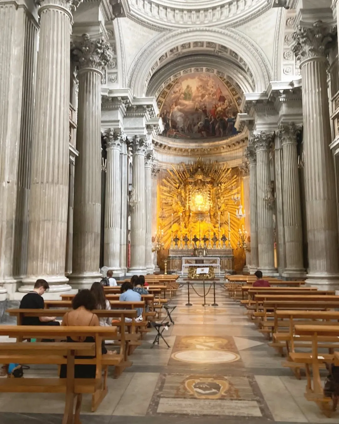 An interior view of a grand cathedral or church, featuring tall columns, rows of wooden pews, and a highly ornate golden altar at the far end. Several individuals are seated in the pews.