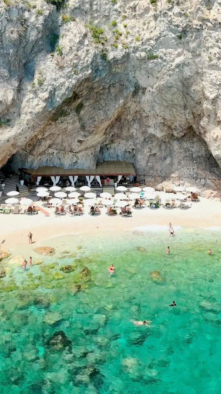 The image depicts a beautiful beach scene with clear turquoise water, a sandy shore, and a unique bar or resort structure built into a large rock formation. People are visible enjoying the water and relaxing on sun loungers under umbrellas.