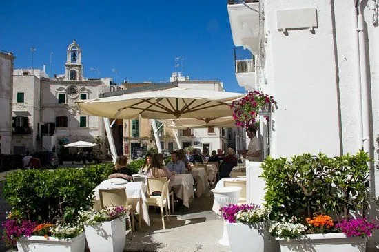 An inviting outdoor dining scene in a Mediterranean-style town square, featuring tables with people under large umbrellas, surrounded by white buildings and vibrant green plants. The sky is clear blue.