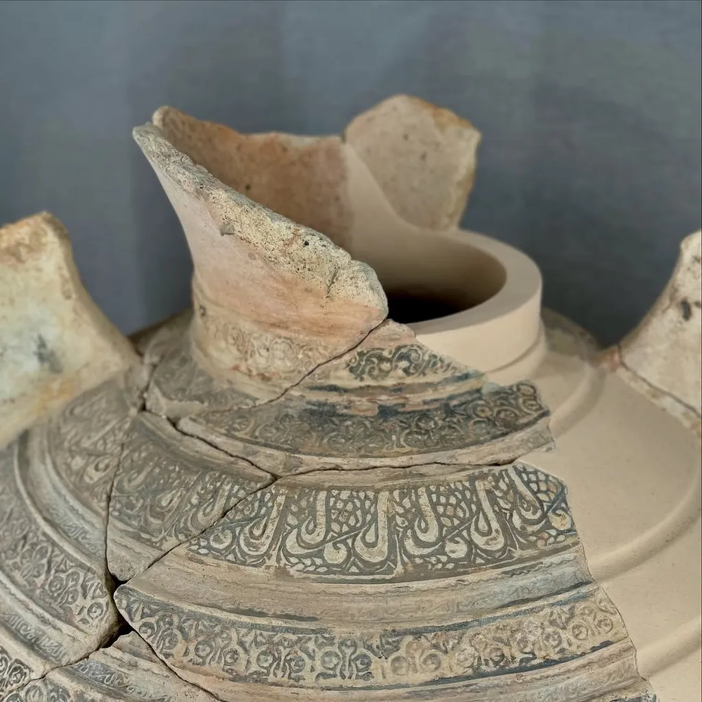 A close-up shot of a broken ancient ceramic pot or vessel, showcasing intricate, faded patterns and visible cracks. The material appears aged and textured.