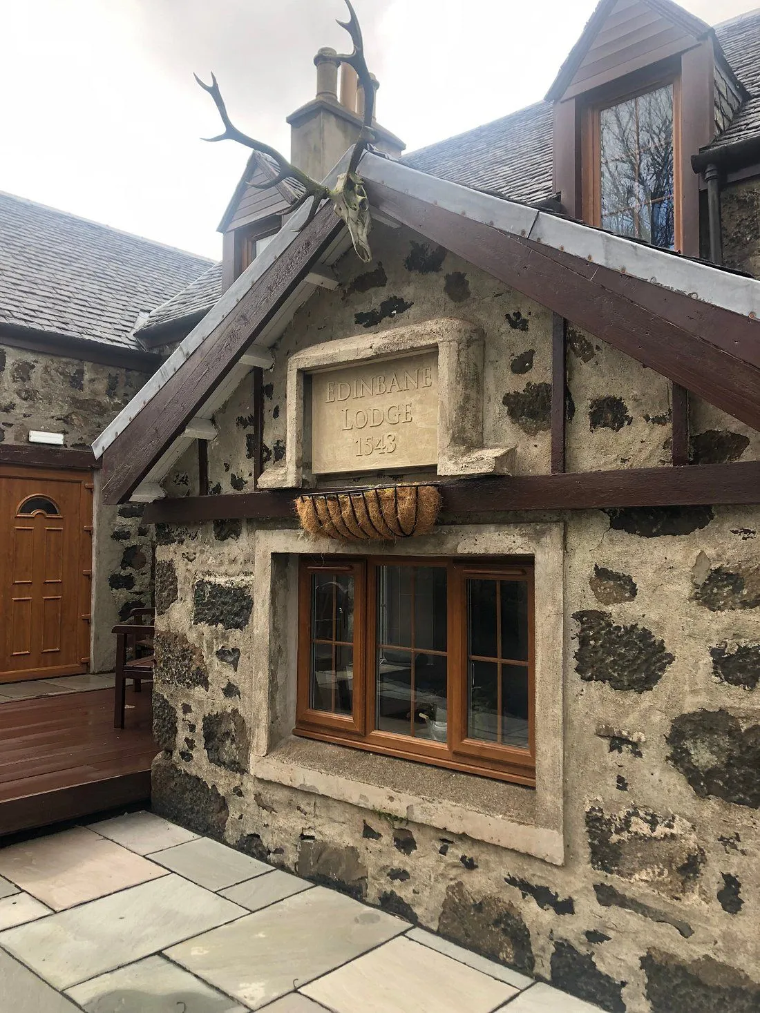 An exterior shot of a stone building, likely a lodge or cottage, featuring a carved stone sign that reads 'EDINBANE LODGE 1543' and a deer skull mounted above it. The building has traditional stone walls and wooden-framed windows. The foreground shows a paved patio area.