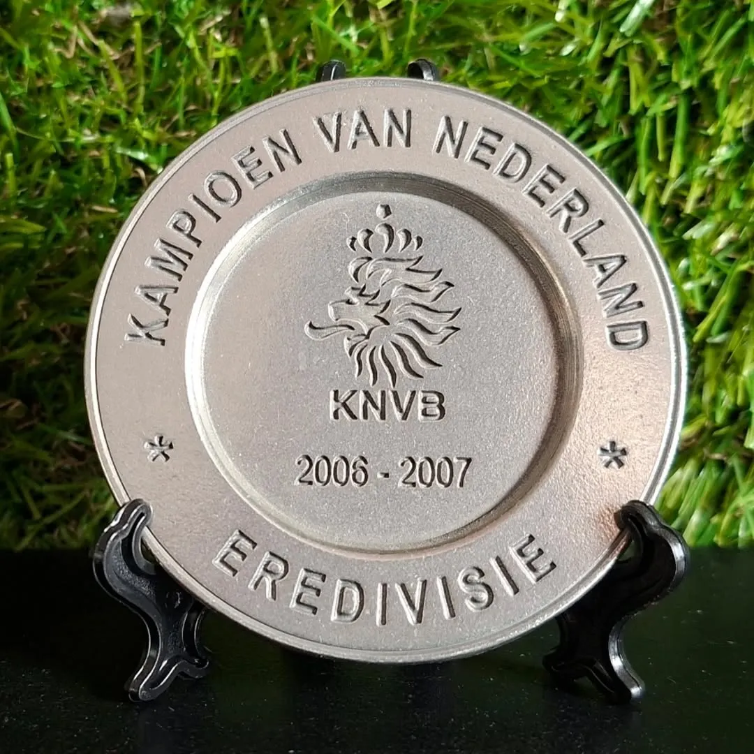 A silver-colored commemorative plate, possibly a medal or trophy, featuring the KNVB logo, the text "KAMPIOEN VAN NEDERLAND", "EREDIVISIE", and "2006-2007". It is displayed on a black stand against a background of artificial green grass.