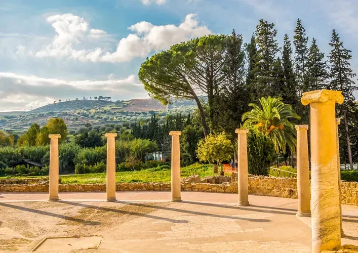 An editorial style image showcasing ancient ruins with several stone pillars in the foreground and a lush green landscape with hills and trees under a partly cloudy sky in the background. The scene is bathed in natural light, casting shadows from the pillars.