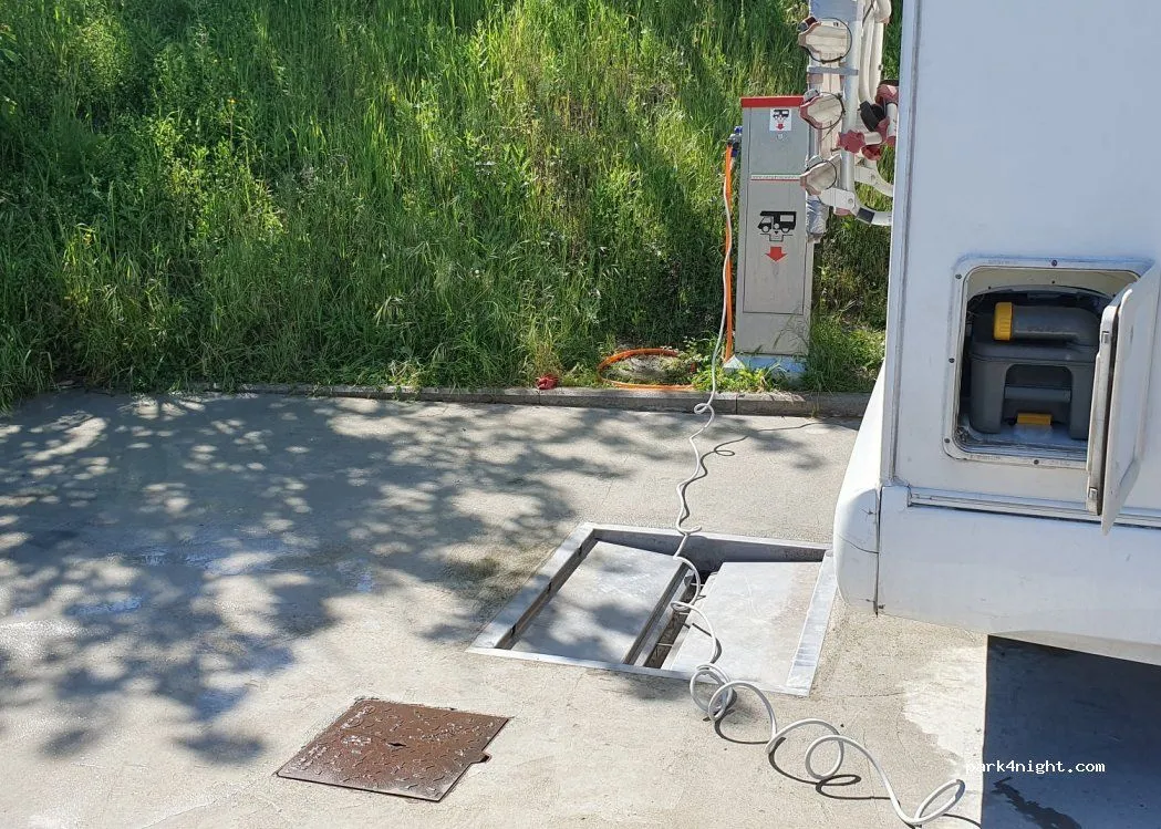 An outdoor scene depicting a service point for a camper or motorhome, with cables connected to a utility post and an open drain on the ground.