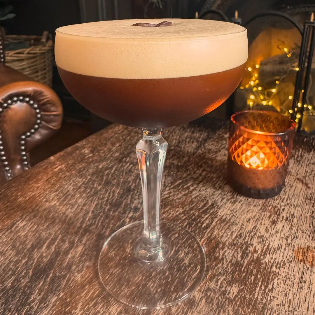 A well-composed and sharp image of an espresso martini, featuring a rich brown liquid topped with creamy foam and garnished with a coffee bean, set in a moody, dimly lit environment.