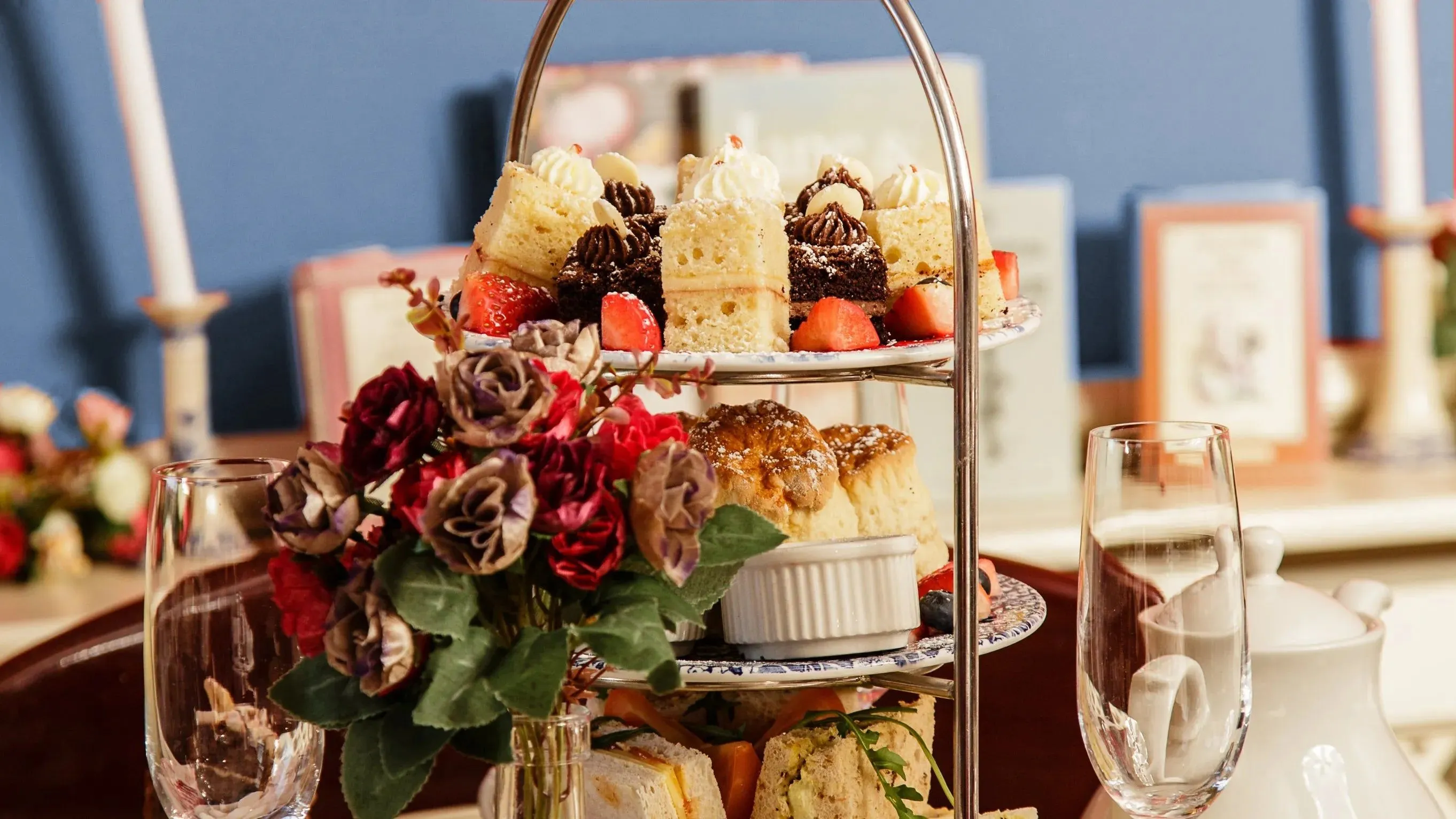 The image features a beautifully arranged tiered tea stand laden with various treats including cakes, scones, and sandwiches, accompanied by a bouquet of flowers. The composition is appealing, highlighting the culinary offerings. The lighting is soft and even, showcasing the textures and details of the food. Most key elements are sharp and in focus, particularly the upper tiers of the stand. Colors appear natural and vibrant, contributing to the photo-realistic quality. The overall presentation is elegant and inviting, fitting for editorial content.
