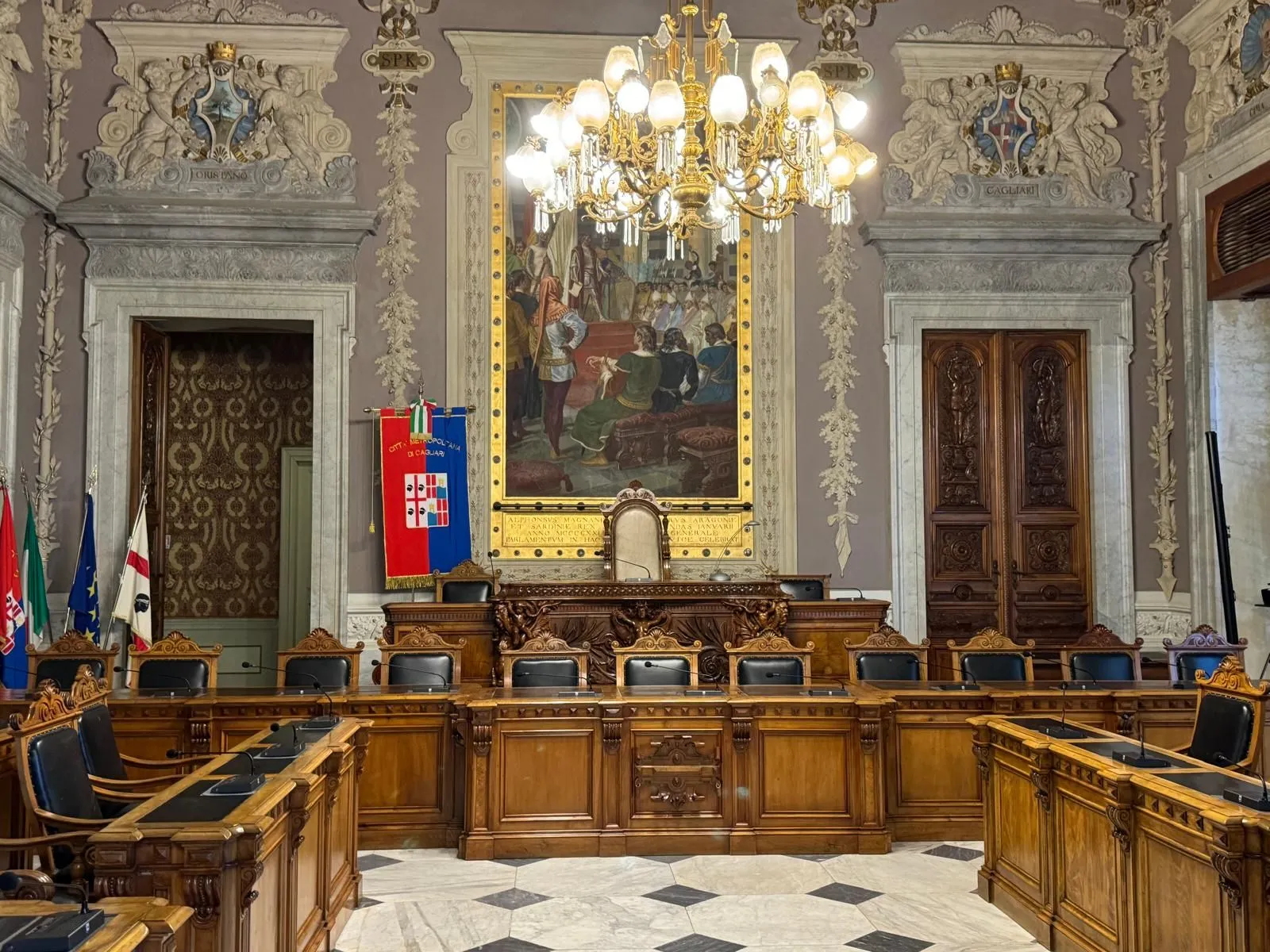 An interior shot of an ornate, historic room, likely a council or meeting hall, featuring a large central painting, a grand chandelier, and dark wooden furniture.