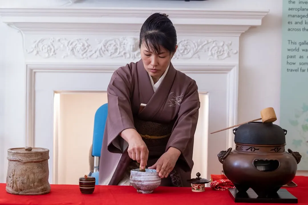 An image depicting a woman in a brown kimono meticulously performing a traditional Japanese tea ceremony. She is focused on whisking matcha in a ceramic bowl, surrounded by various tea ceremony utensils on a red table. A white, ornate fireplace mantel serves as the background.
