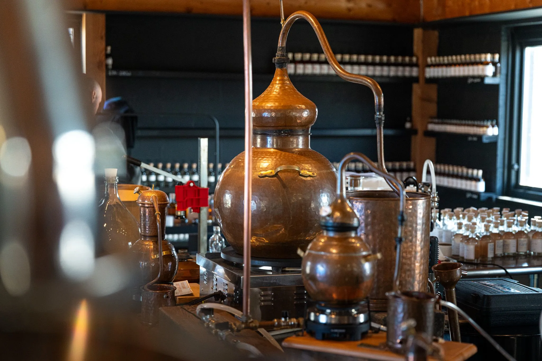A clear photograph of copper stills and distillation equipment, likely in a distillery setting, with bottles visible on shelves in the background.