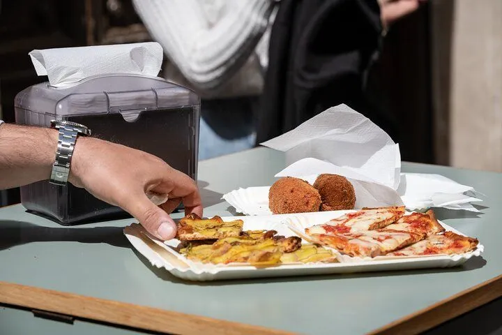 A close-up shot captures a variety of Italian street food, including slices of pizza and arancini, served on disposable paper plates. A hand reaches for a piece of food, with a napkin dispenser partially visible on the left. The scene suggests a casual dining experience.