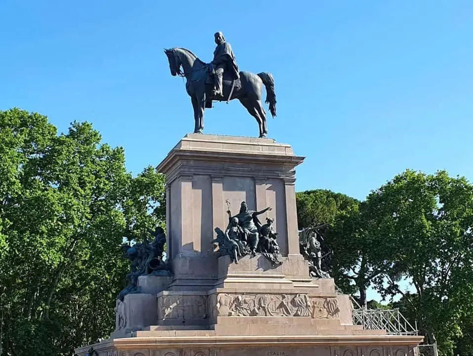 An outdoor photograph of a large bronze equestrian statue on a stone pedestal, set against a clear blue sky and green trees.