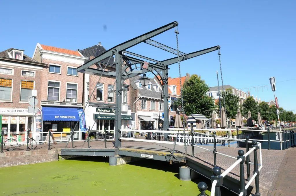 An image featuring a traditional drawbridge over a canal, surrounded by old-fashioned buildings and shops under a clear blue sky. The canal water appears green, indicating algae growth, which is a common sight in such waterways.