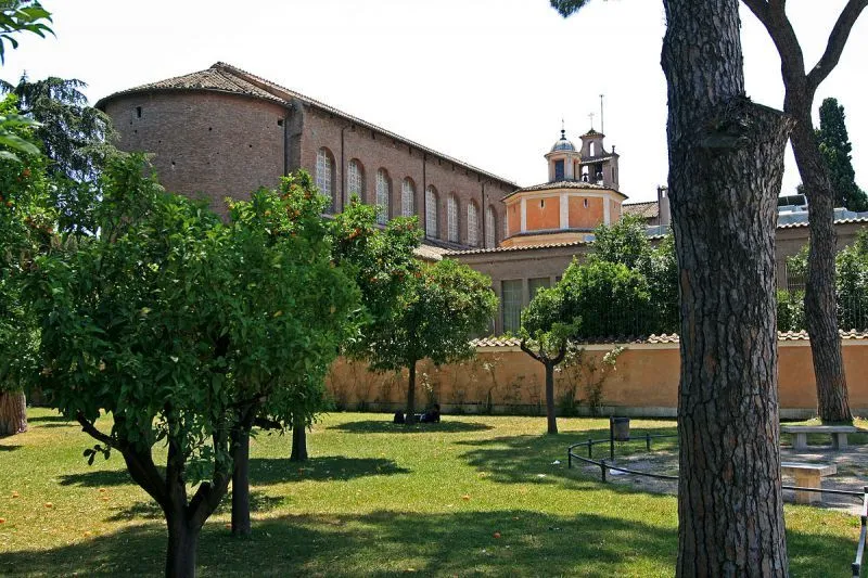An exterior view of a historic brick building with arched windows and a dome, framed by green trees and a grassy lawn under a sunny sky. The image captures an architectural landmark in a garden setting.